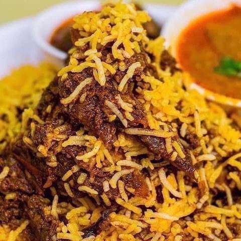 Close-up of yellow briyani rice over dark mutton pieces, a bowl of curry behind