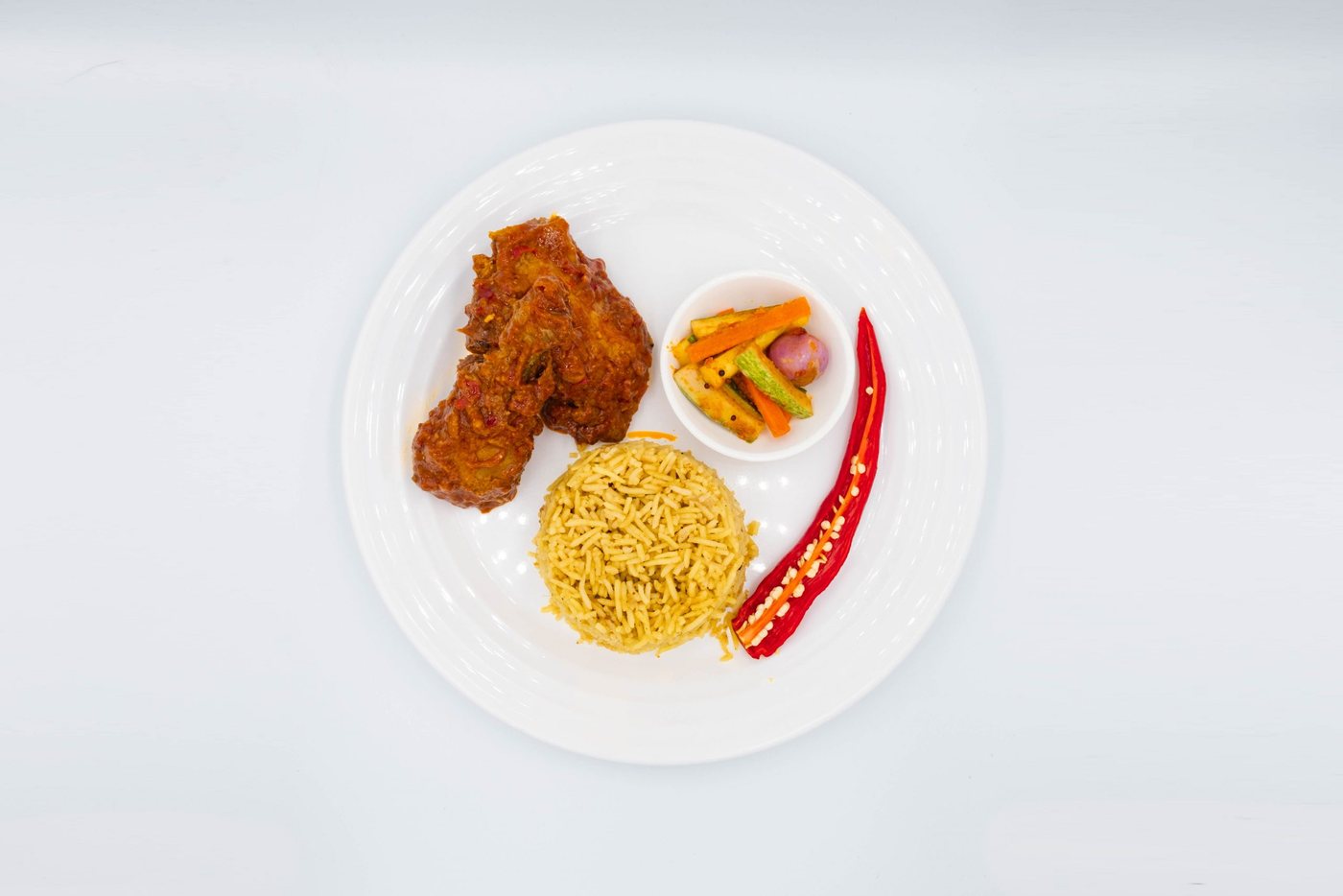 Plate of yellow rice, chicken in red sauce, pickle cup and a halved red chilli, seen from above